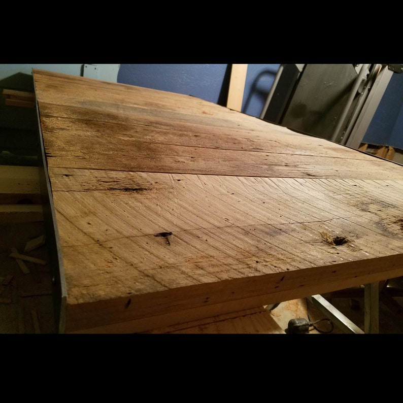 Puede incluir: Una gran mesa rectangular de madera recuperada. La madera tiene un aspecto natural y envejecido con vetas, nudos e imperfecciones visibles. La mesa est&aacute; construida con varias tablas de madera, creando una superficie r&uacute;stica y texturizada.