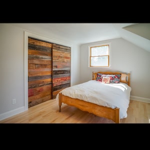 Puede incluir: Dormitorio con una cama de madera y puertas correderas de armario hechas de madera reciclada. Las puertas est&aacute;n compuestas por tablas de madera de varios colores. La cama tiene un edred&oacute;n blanco y almohadas decorativas. Se ve una ventana.