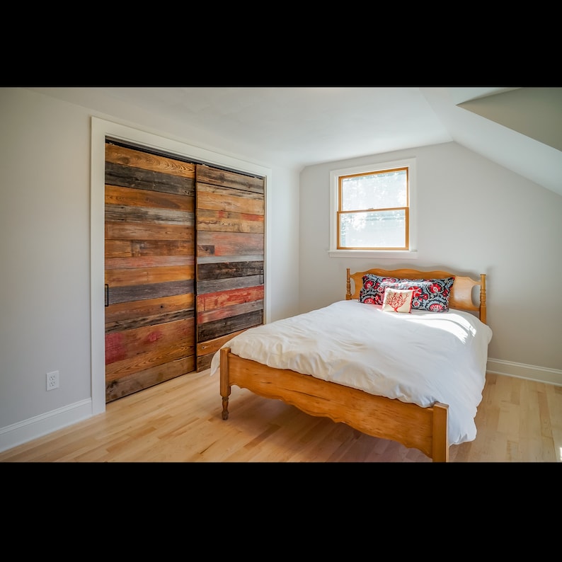 Puede incluir: Dormitorio con una cama de madera y puertas correderas de armario hechas de madera reciclada. Las puertas est&aacute;n compuestas por tablas de madera de varios colores. La cama tiene un edred&oacute;n blanco y almohadas decorativas. Se ve una ventana.