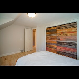 Puede incluir: Interior de dormitorio con una cama blanca, suelo de madera clara y un armario corredero r&uacute;stico con puerta de granero. La puerta del armario es de madera reciclada en tonos marr&oacute;n, rojo y naranja. Una puerta blanca conduce a otra habitaci&oacute;n.