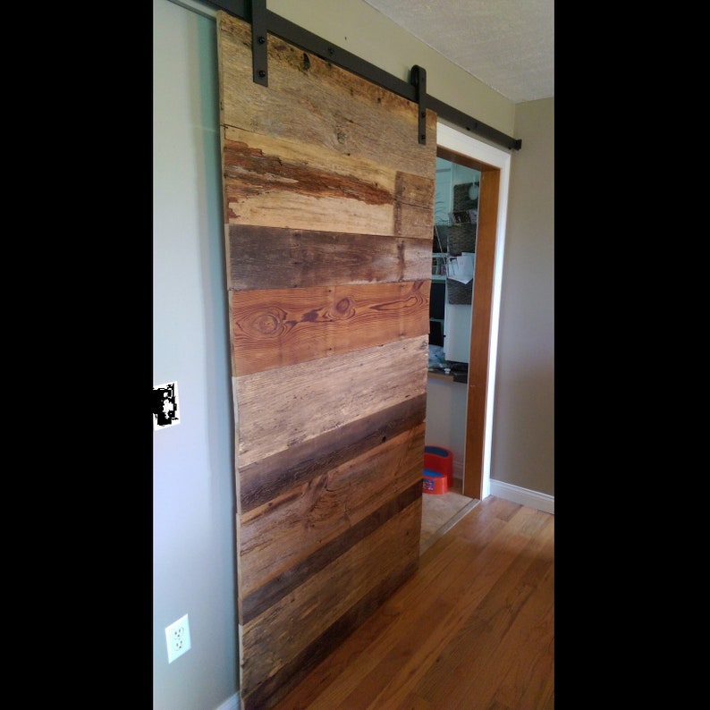 Puede incluir: Una puerta de granero r&uacute;stica hecha de madera reciclada, con una mezcla de tonos marrones, beige y grises. La puerta est&aacute; montada sobre un riel corredizo de metal negro, agregando un toque industrial al espacio interior.
