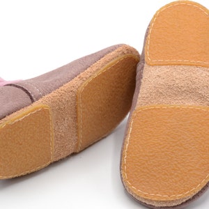 Könnte beinhalten: Ein Paar weiche, rosa und braune Leder-Babyschuhe mit einem Obermaterial aus Wildleder und einer strukturierten Gummisohle.