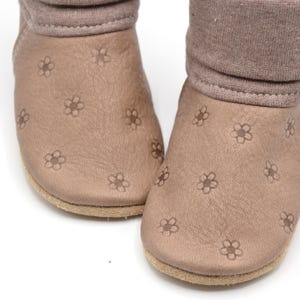 Könnte beinhalten: Ein Paar braune Leder-Babyschuhe mit Blumenmuster. Die Schuhe haben einen umgeschlagenen Bund und ein weiches, bequemes Futter.