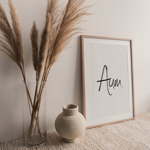 May include: A framed print with the word "Aum" written in black calligraphy on a white background. The print is sitting on a beige woven rug with a vase of dried pampas grass and a small beige ceramic vase.