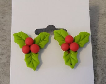 May include: A pair of green and red holly berry stud earrings. The earrings are made of polymer clay and feature a cluster of red berries with green leaves.