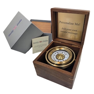 May include: A brass gimbal compass housed in a square dark wood box. The interior lid features a gold-colored plate with the text "Personalize Me! Add your own unique message". A grey box and a small card with "Stanley London" text are included.