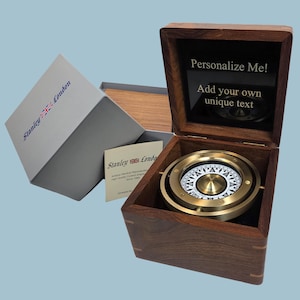 Peut inclure: Une boussole nautique en laiton logée dans une boîte en bois foncé avec un couvercle à charnière. L'intérieur du couvercle porte le texte "Personalize Me! Add your own unique text". Une boîte grise et une carte avec "Stanley London" sont présentes.