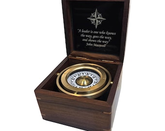 May include: A dark wood box containing a brass gimbaled compass. The interior lid features a compass rose icon and the engraved text: A leader is one who knows the way, goes the way, and shows the way. John Maxwell. The compass has a white dial.