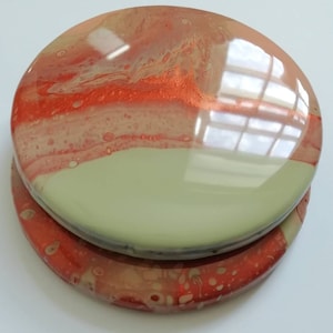May include: Two round coasters with a glossy finish. The top coaster features a blend of red, orange, and white hues, while the bottom coaster has a similar color scheme. The coasters have a unique, marbled appearance.