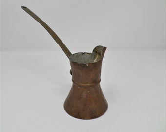 Pode incluir: Uma cafeteira de cobre com uma alça de latão. A cafeteira tem uma base arredondada e um bico alargado. A cafeteira está ligeiramente desbotada.