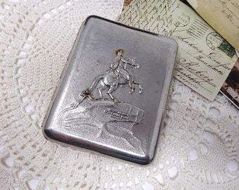 May include: Silver cigarette case with an embossed design of a person on horseback. The case has a worn, vintage look.