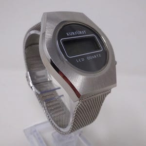 May include: A silver-toned wristwatch with a black LCD display. The watch face reads "Kurf&uuml;rst LCD Quartz". The watch has a silver metal mesh band.