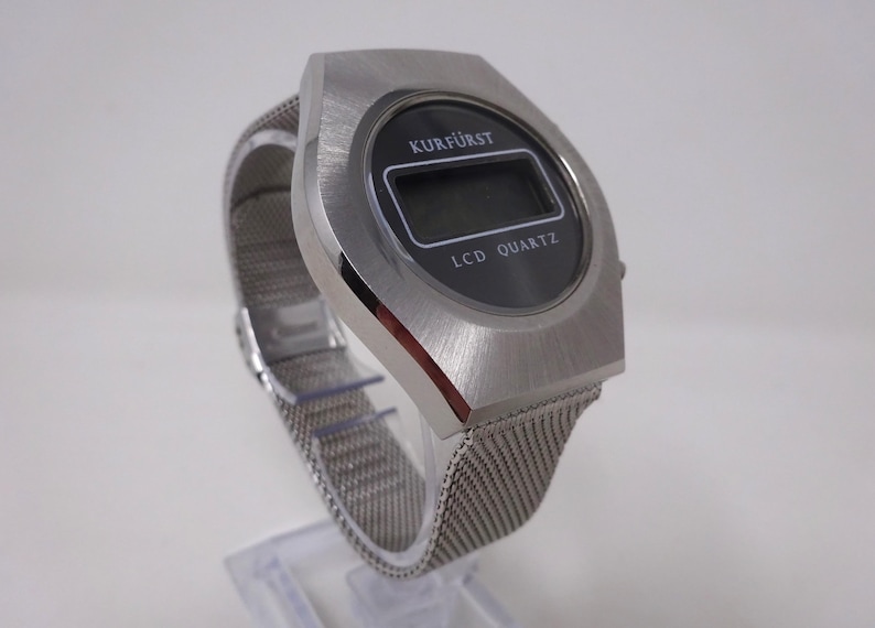 May include: A silver-toned wristwatch with a black LCD display. The watch face reads "Kurf&uuml;rst LCD Quartz". The watch has a silver metal mesh band.