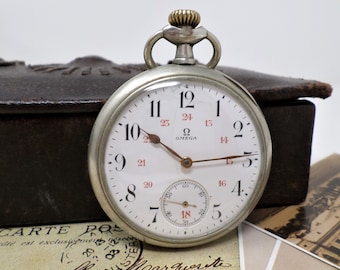 May include: A silver pocket watch with a white face and black hands. The watch has a 24-hour dial and the text "Ω OMEGA" printed on the face.
