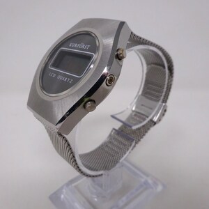 May include: A silver digital watch with a black face and a silver mesh band. The watch face displays the text "KURF&Uuml;RST" and "LCD QUARTZ".