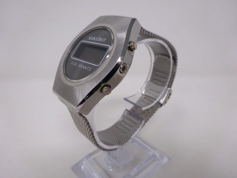 May include: A silver digital watch with a black face and a silver mesh band. The watch face displays the text "KURF&Uuml;RST" and "LCD QUARTZ".
