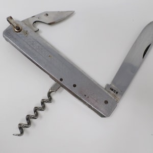Pode incluir: Uma multi-ferramenta vintage com acabamento em aço inoxidável. Inclui um abridor de garrafas, um saca-rolhas e uma lâmina de faca dobrável. A lâmina é marcada com a palavra "INOX".