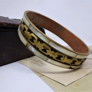 May include: A gold and white bangle bracelet with a decorative design. The bracelet is made of brass and features a white mother-of-pearl inlay.