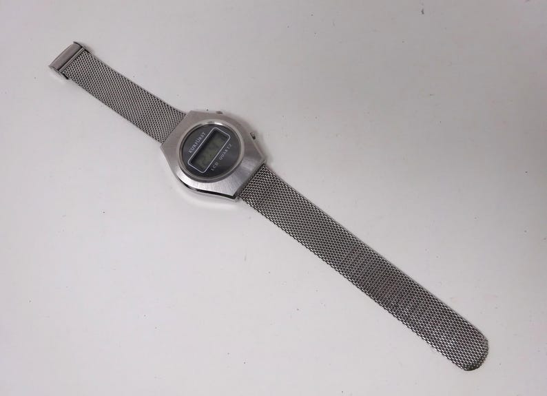 May include: A silver digital watch with a mesh metal band. The watch face displays the time and date.