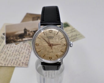 May include: A silver-toned wristwatch with a black leather band. The watch face is cream-colored with black markings and the text "SINEX GENEVE 17 Jewels SWISS MADE" printed on it.