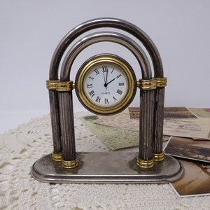 Può includere: Un orologio da tavolo in metallo argentato con quadrante circolare, numeri romani neri e dettagli dorati. L'orologio presenta una cornice ad arco sostenuta da due colonne scanalate su una base ovale. La parola QUARTZ è stampata sul quadrante bianco.