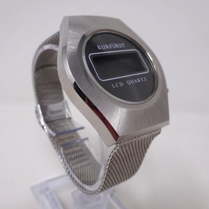 May include: A silver-toned wristwatch with a black digital display. The watch face reads "KURF&Uuml;RST LCD QUARTZ". The watch has a silver mesh band.
