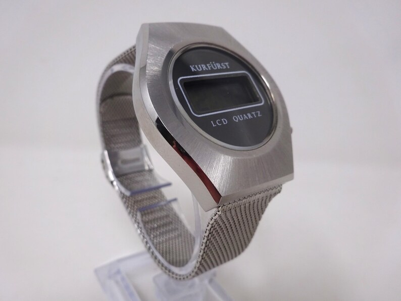 May include: A silver-toned wristwatch with a black digital display. The watch face reads "KURF&Uuml;RST LCD QUARTZ". The watch has a silver mesh band.