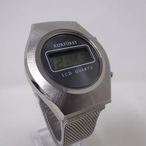 May include: A silver digital watch with a black face and a mesh band. The watch face displays the time "12:48" and the text "KURF&Uuml;RST LCD QUARTZ".