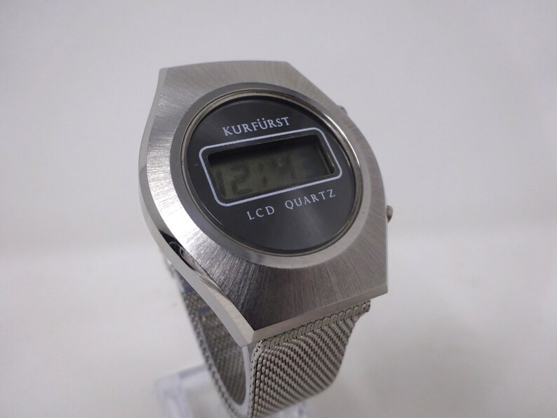 May include: A silver digital watch with a black face and a mesh band. The watch face displays the time "12:48" and the text "KURF&Uuml;RST LCD QUARTZ".