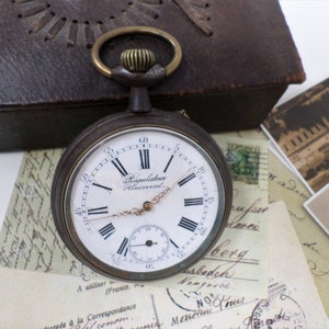 May include: A vintage silver pocket watch with Roman numeral markings on the face. The watch face reads "Régulateur Universel".