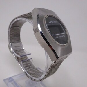 May include: A silver-toned digital wristwatch with a black face and a mesh metal band. The watch face displays "KURFURST" and "LCD QUARTZ".