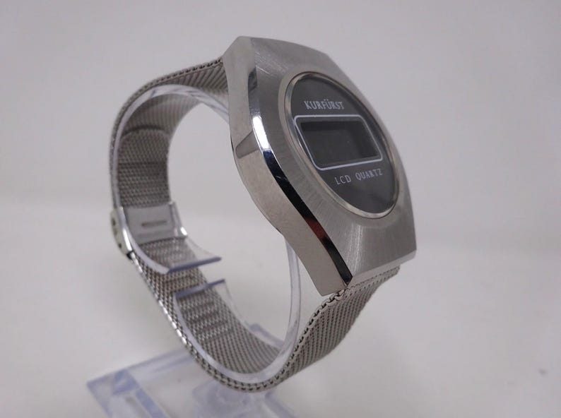 May include: A silver-toned digital wristwatch with a black face and a mesh metal band. The watch face displays "KURFURST" and "LCD QUARTZ".