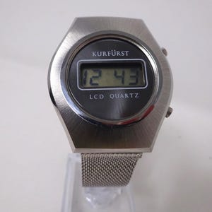 May include: A silver-toned digital wristwatch with a black face and a mesh metal band. The watch face displays the time "12:43" and the text "KURF&Uuml;RST LCD QUARTZ".