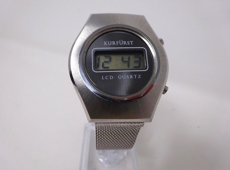 May include: A silver-toned digital wristwatch with a black face and a mesh metal band. The watch face displays the time "12:43" and the text "KURF&Uuml;RST LCD QUARTZ".