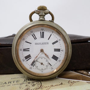May include: A silver pocket watch with Roman numeral markings on the face. The watch face is white with black markings and the word "REGULATEUR" printed in black. The watch has a gold-colored trim around the edge of the face.