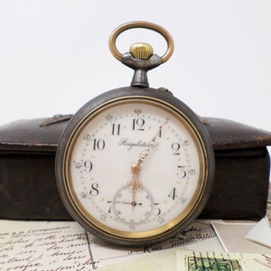 May include: A vintage silver pocket watch with a white face and black Roman numeral hour markers. The watch has a gold-colored bezel and a gold-colored winding stem. The watch face also has the word "Regulateur" printed on it.