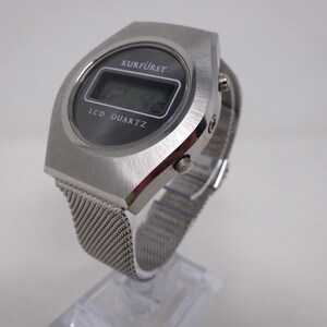 May include: A silver-toned digital wristwatch with a black face and a mesh metal band. The watch face displays the text "KURF&Uuml;RST" and "LCD QUARTZ".