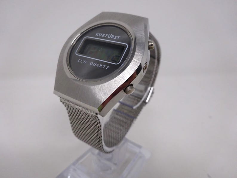 May include: A silver-toned digital wristwatch with a black face and a mesh metal band. The watch face displays the text "KURF&Uuml;RST" and "LCD QUARTZ".