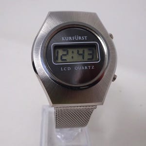 May include: A silver-toned digital wristwatch with a black face and a mesh metal band. The watch face displays the time as 12:43 and the words "KURF&Uuml;RST LCD QUARTZ".