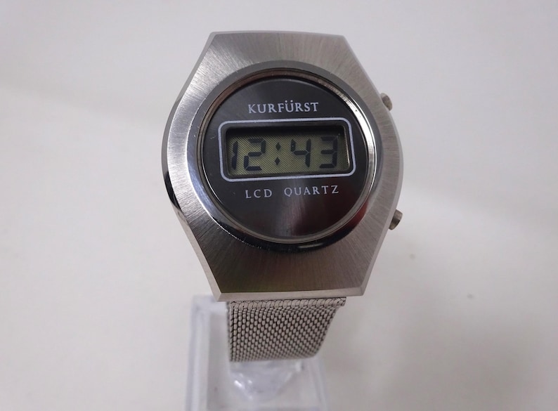 May include: A silver-toned digital wristwatch with a black face and a mesh metal band. The watch face displays the time as 12:43 and the words "KURF&Uuml;RST LCD QUARTZ".