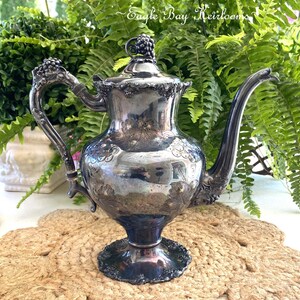 May include: A silver teapot with an ornate design of grapes and leaves. The teapot has a round body and a curved spout. It sits on a woven mat.