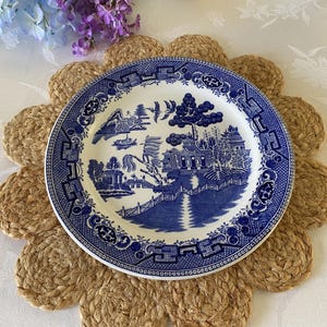 May include: A blue and white ceramic plate with a landscape design rests on a woven jute placemat. The placemat is shaped like a flower with a natural tan color. The plate features a detailed illustration of a bridge, buildings, and trees.