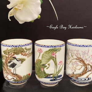 May include: Three white ceramic cups with hand-painted Japanese-style designs. Each cup features a different nature scene: a crane, lotus flowers, and cherry blossoms. The cups have blue and gold trim.