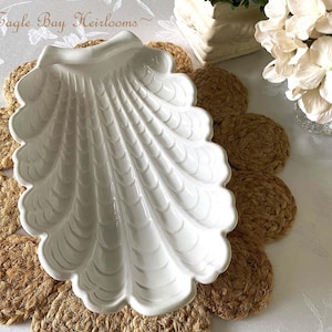 May include: A white, shell-shaped serving dish with a scalloped edge. The dish is placed on a woven, circular placemat. The text "Eagle Bay Heirlooms" is visible in the image.