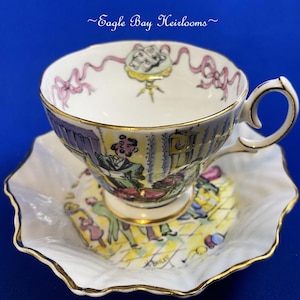 May include: A vintage teacup and saucer set with a whimsical illustration. The white porcelain cup and saucer feature a colorful cartoon scene with gold trim. The cup has a curved handle and the saucer has a scalloped edge.
