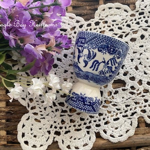 May include: A blue and white ceramic egg cup with a detailed Willow pattern. The cup is set on a white crocheted doily, with purple and white flowers in the background. The text "Eagle Bay Heirlooms" is visible.