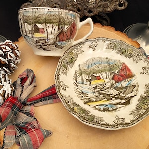 May include: A vintage teacup and saucer set with a winter landscape scene featuring red barns and a snowy river. The cup and saucer have a white background with a detailed illustration. The set is placed on a wooden surface with decorative pinecones and a plaid ribbon.