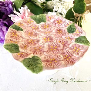 May include: A ceramic plate with a floral design. The plate is shaped like a cluster of pink flowers with yellow centers and green leaves. The plate has a slightly raised, textured surface. The text "Eagle Bay Heirlooms" is at the bottom.