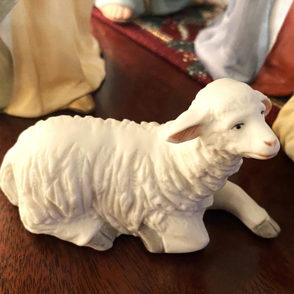 Może przedstawiać: Biała porcelanowa figurka leżącego baranka z głową zwróconą w bok. Baranek ma szczegółowe, fakturowane futro i słodki wyraz twarzy.