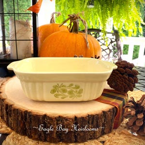 May include: Cream-colored rectangular baking dish with green floral design, resting on a wooden slice. The dish is part of a fall-themed display with pumpkins, pinecones, and a lantern. The text "Eagle Bay Heirlooms" is visible.